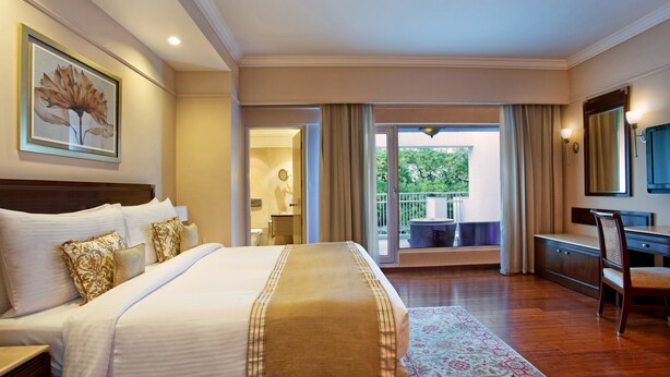 A hotel room with a large bed, a desk, a chair, a bathroom, and a balcony with a view of greenery. - The Claridges, New Delhi (New Delhi)