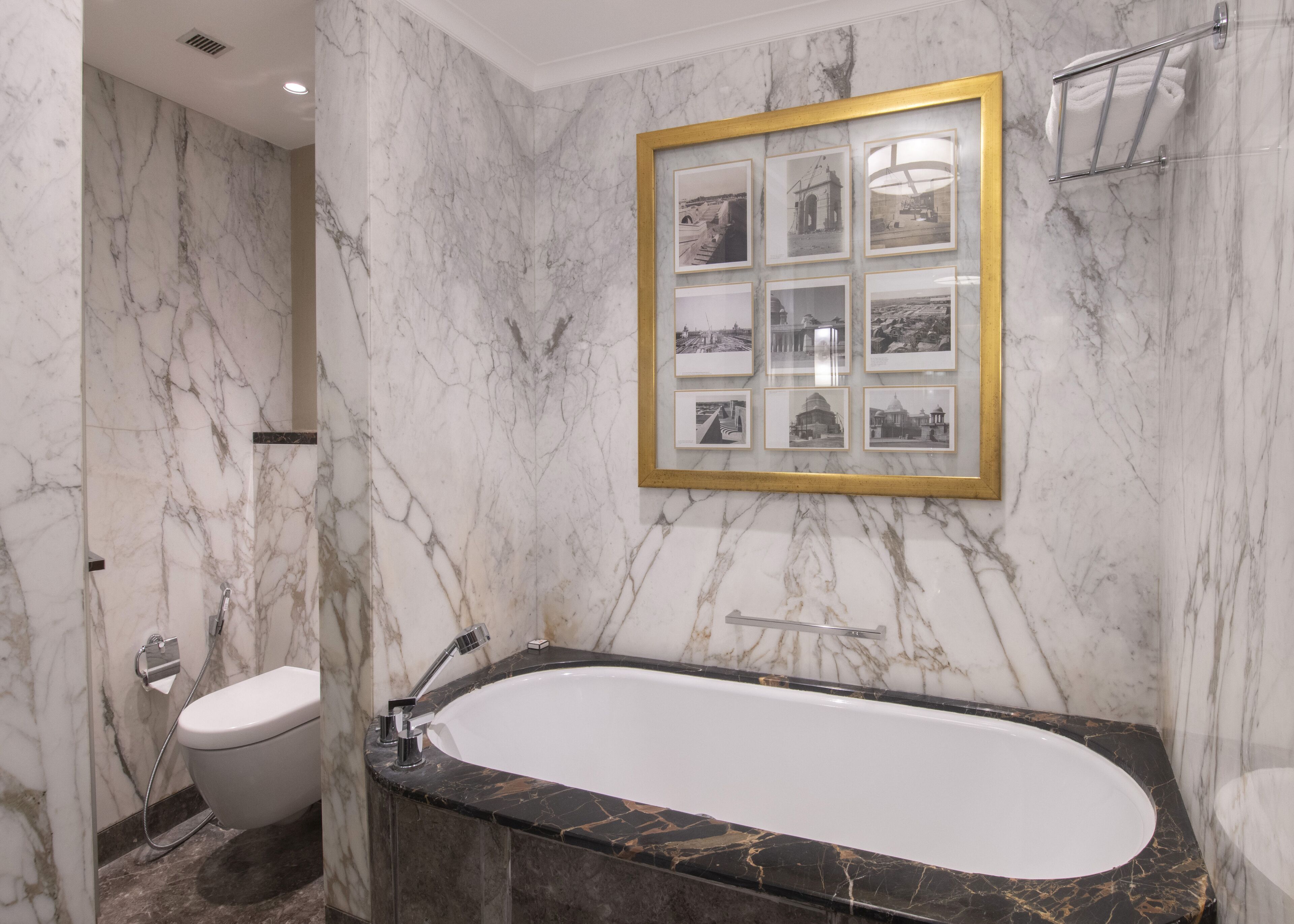 Un baño con pared de mármol, una bañera, un inodoro y un cuadro enmarcado con una vista urbana.