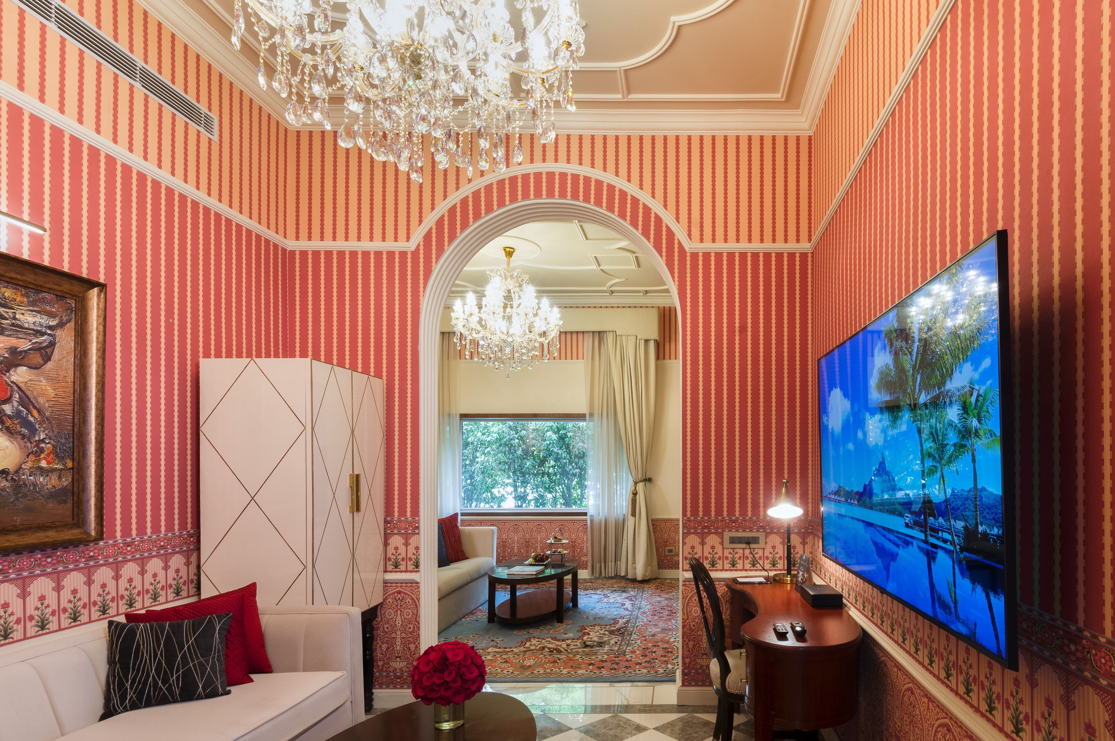 Una habitación con papel tapiz a rayas, un candelabro, un sofá, un televisor y una ventana con vistas a árboles.