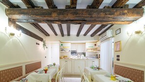 A dining area with tables set for a meal, a kitchen in the background, and a ceiling with exposed wooden beams. - Locanda Ca' Zose (Venice)