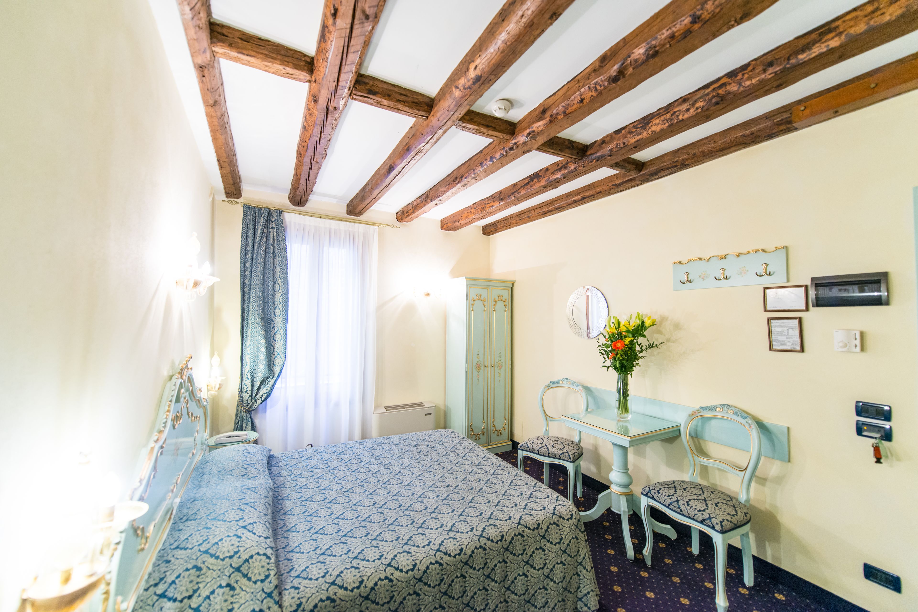 A bedroom with a bed, a vanity with a mirror, a chair, and a table with a vase of flowers.