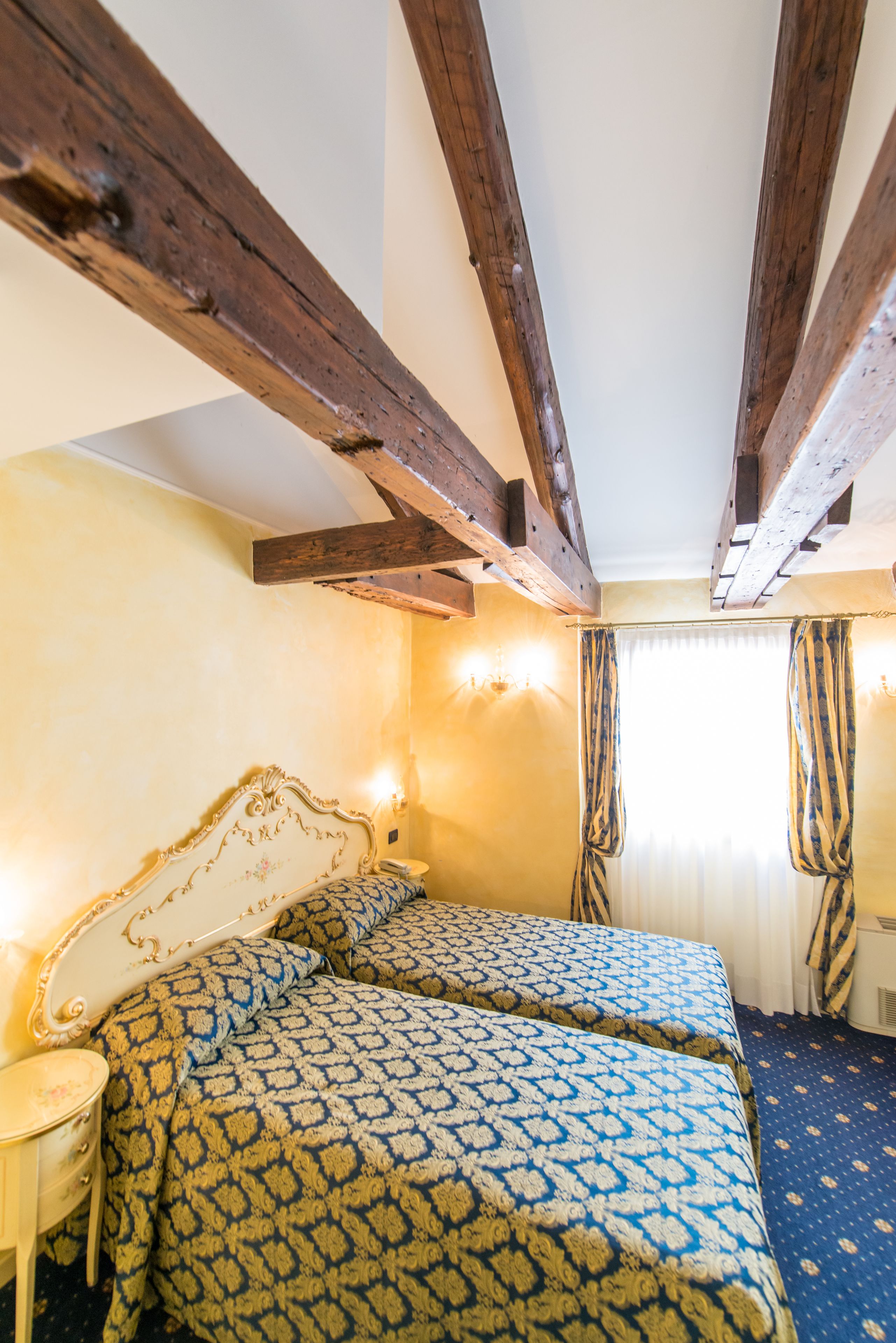 A bedroom with two beds, wooden beams, a patterned bedspread, and a window with curtains.