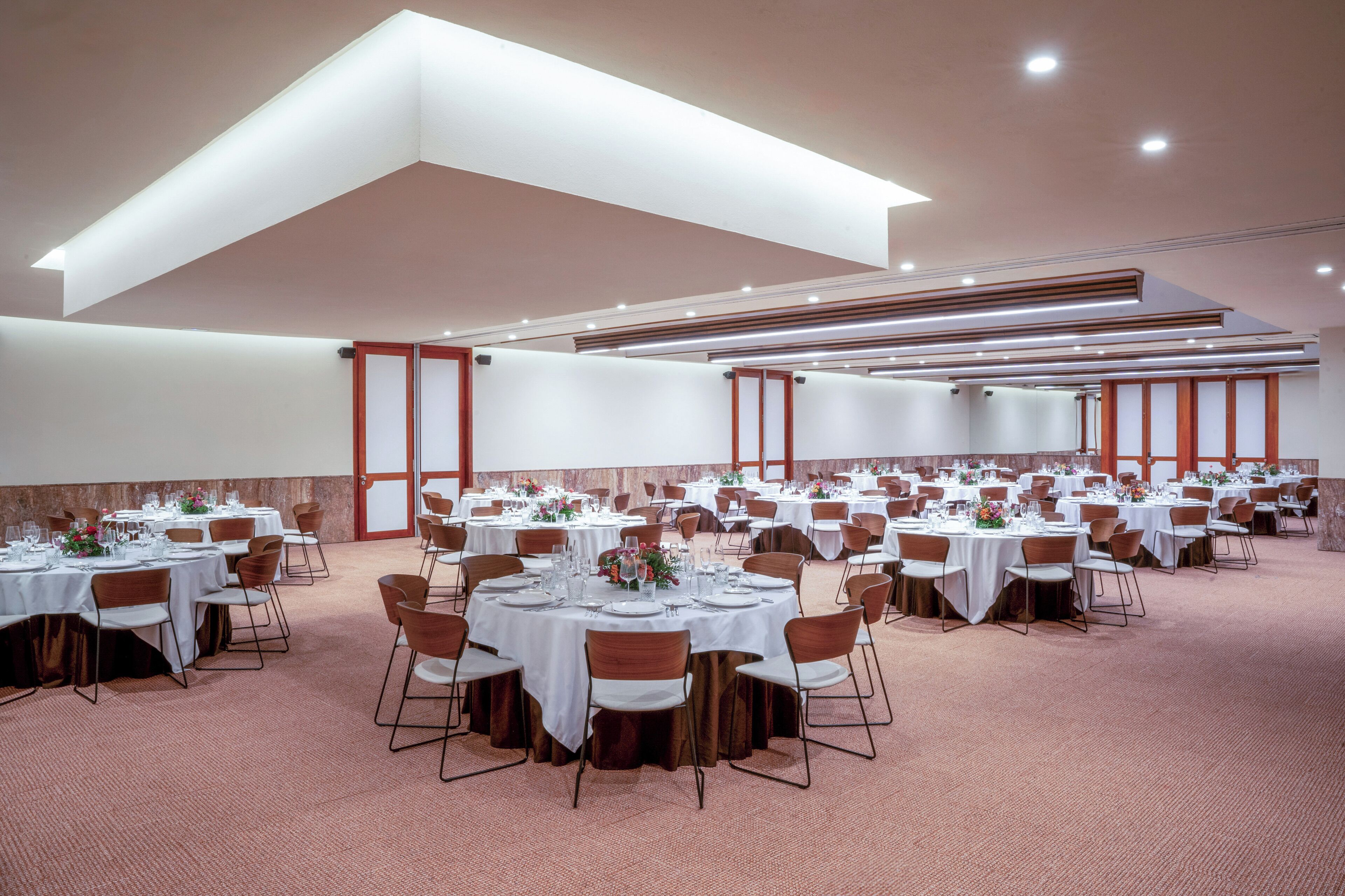 A large banquet hall with multiple round tables, each set with white tablecloths, chairs, and floral centerpieces.