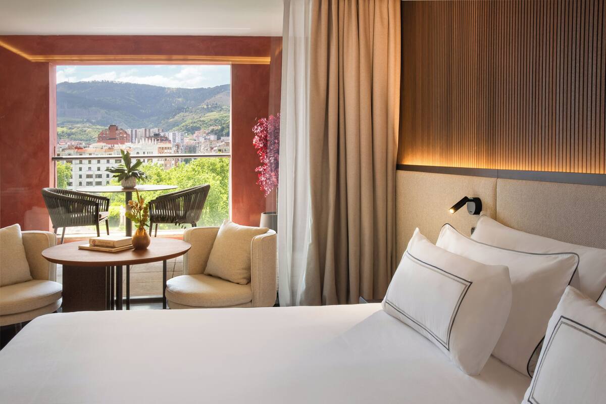 A hotel room with a large bed, two armchairs, a small table with a book and a vase, and a view of a cityscape and mountains through a large window.