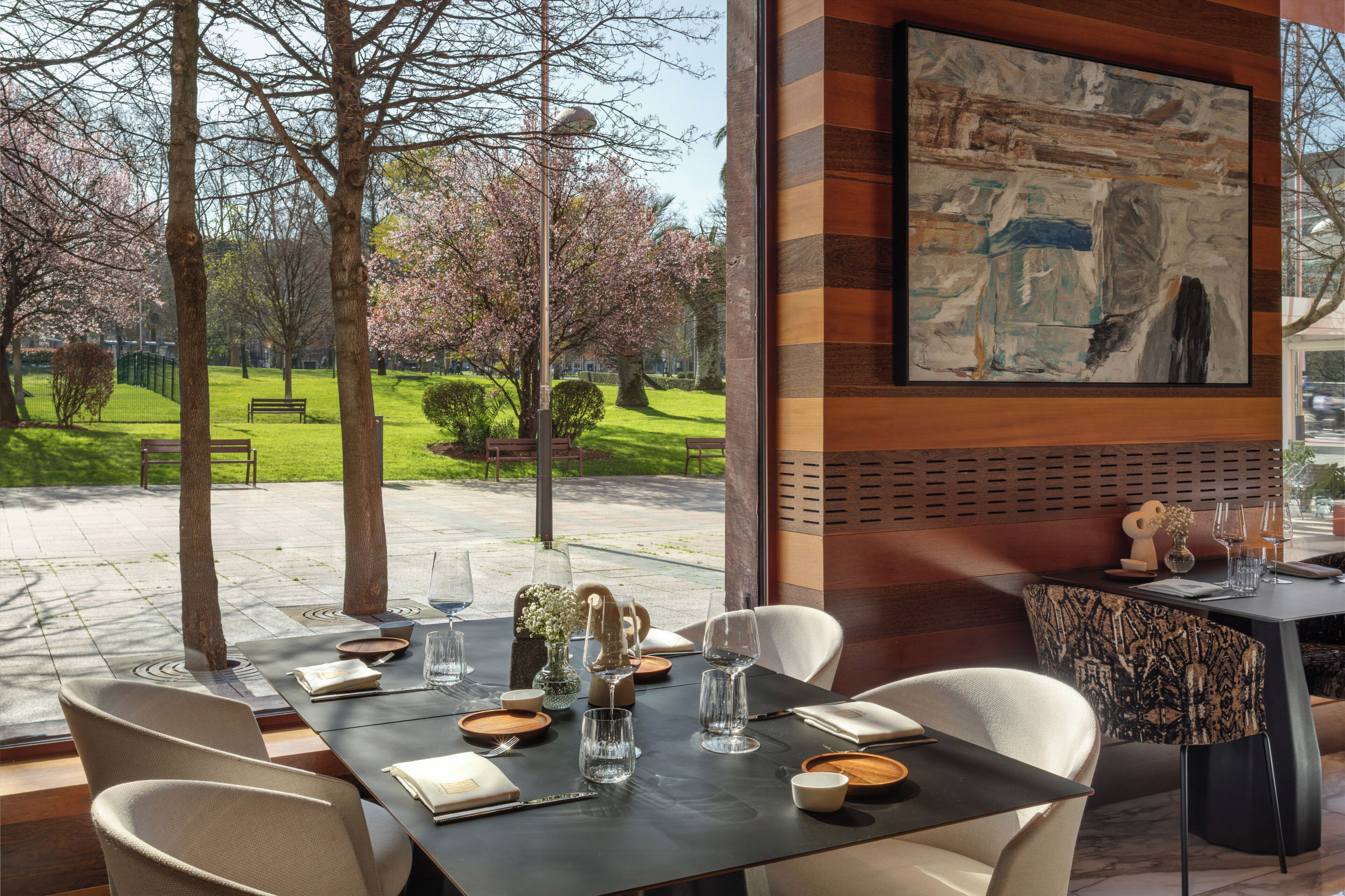 Un espacio de comedor al aire libre con una mesa dispuesta para dos, un banco y vistas a un parque con árboles y césped.