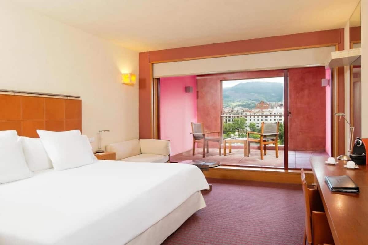 A hotel room with a large bed, a desk, a chair, and a view of a cityscape through a sliding door.