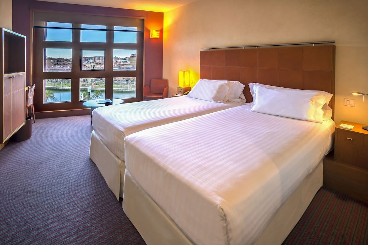 A hotel room with two beds, a television, a desk, and a view of a cityscape through large windows.