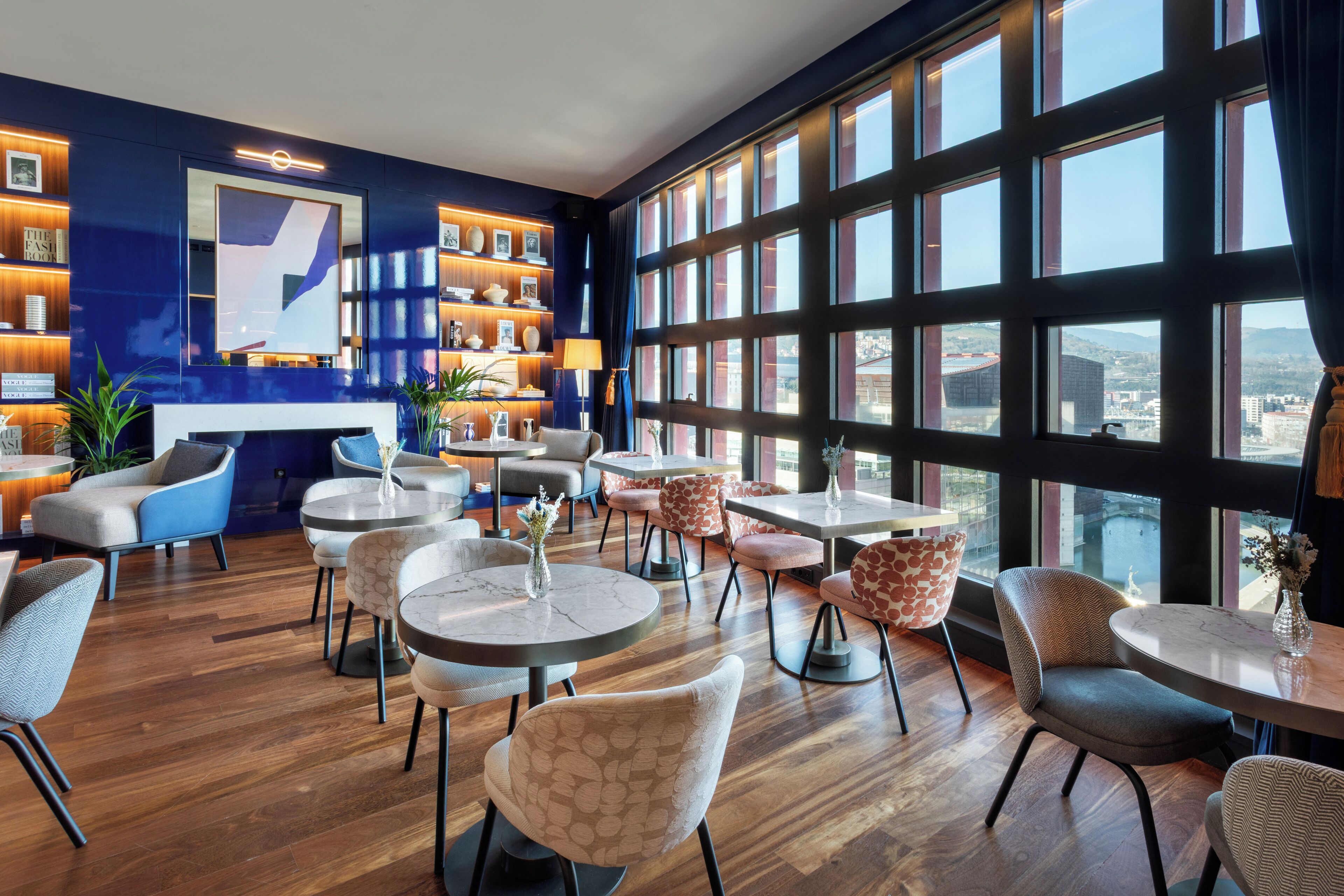 A modern interior with round tables and chairs, a large window with a city view, and a bookshelf with decorative items.