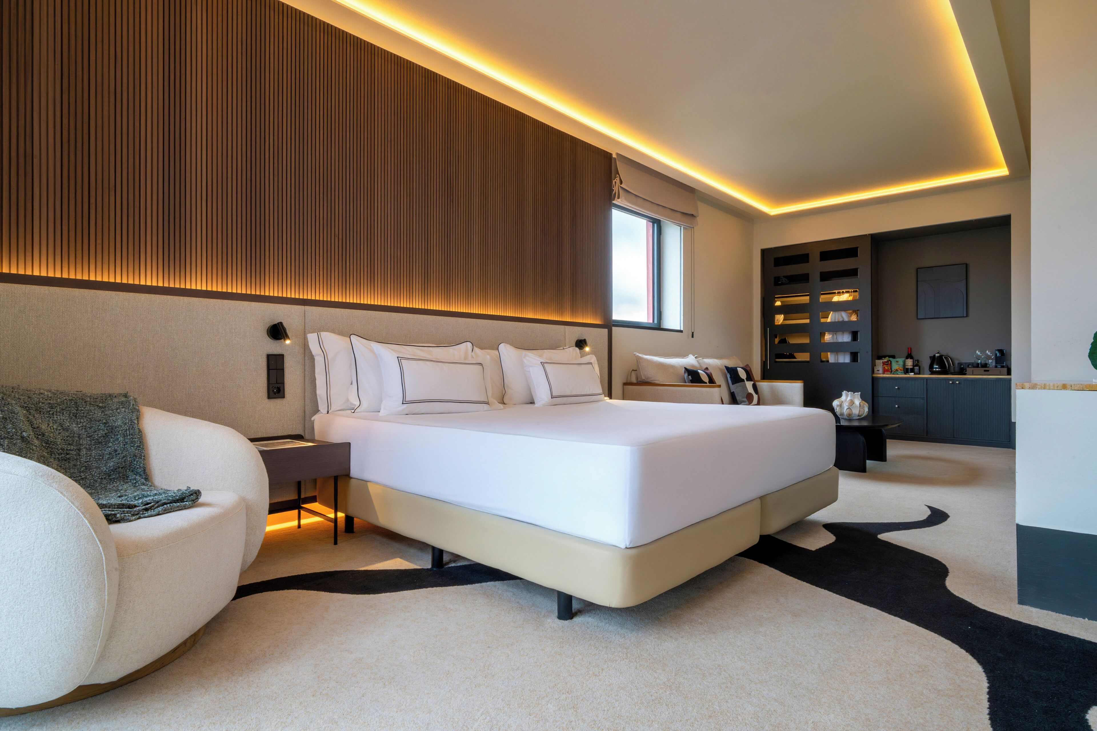 A modern bedroom with a large bed, a sofa, a nightstand, and a built-in shelving unit with various items.