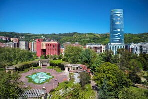 Panorama urbano con un moderno grattacielo in evidenza, una fontana storica e vegetazione circostante.