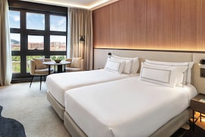 Camera d'albergo con due letti, un tavolino rotondo e vista sui palazzi attraverso ampie finestre.
