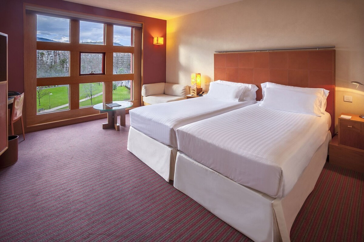 Habitación de hotel con dos camas, un sofá, una mesita y ventana con vistas a un paisaje verde.