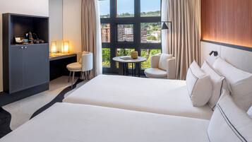 Habitación de hotel con una cama grande, un escritorio con silla, una mesita y vistas a edificios a través de amplias ventanas.