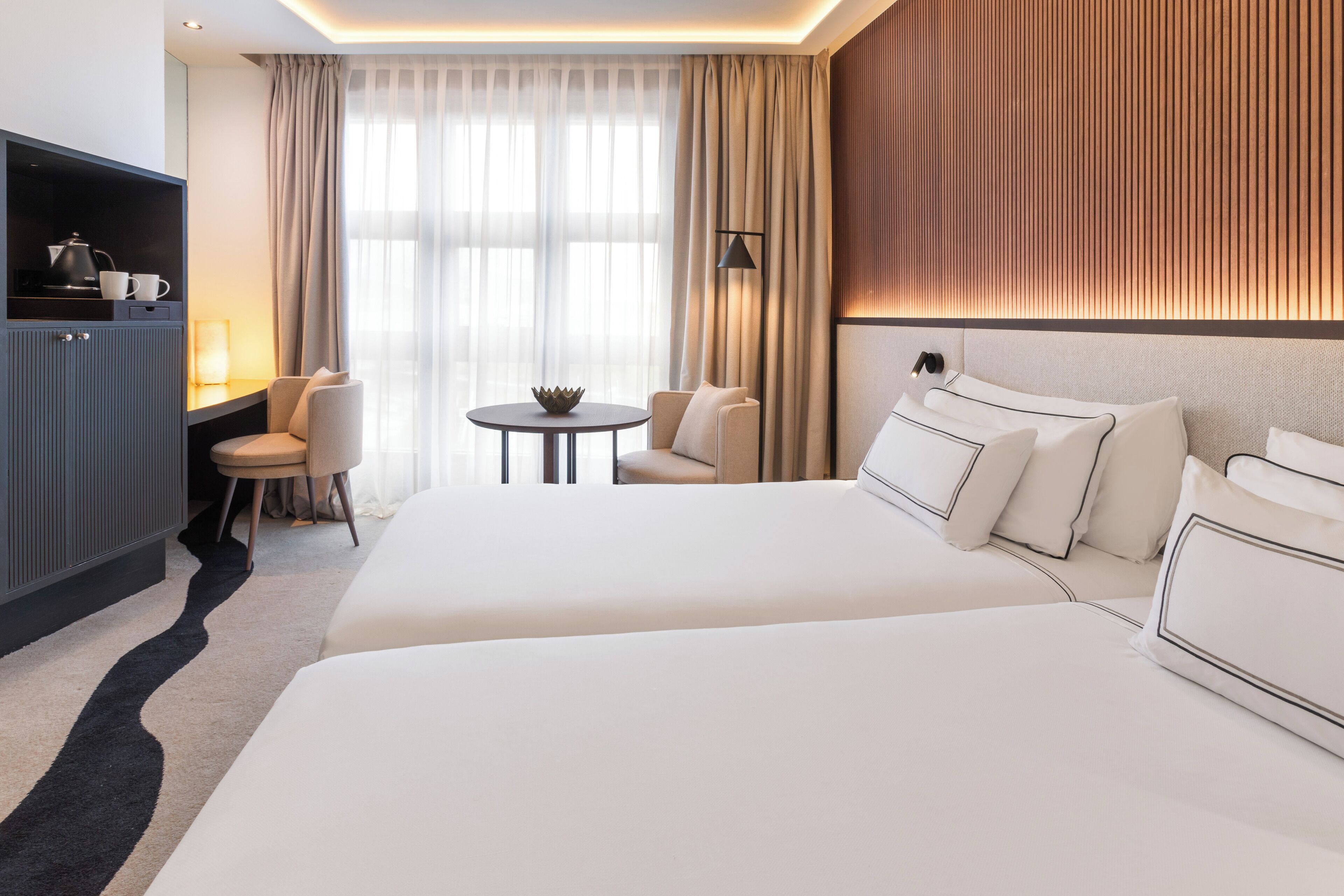 Habitación de hotel con una cama grande, un escritorio con hervidor, una silla y una mesita con un bol.
