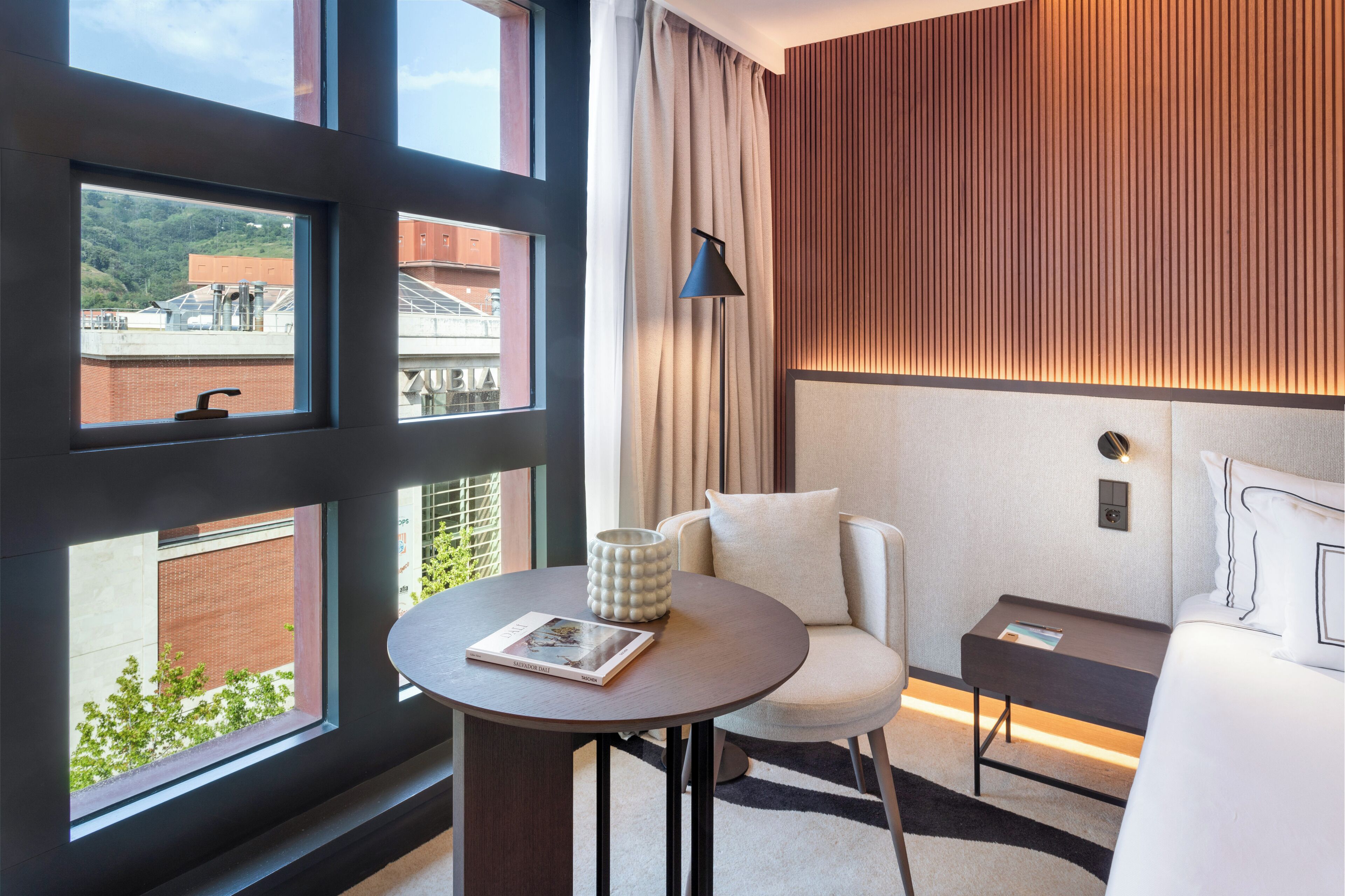 Una habitación de hotel moderna con un ventanal grande, una mesa redonda, un sofá y una cama.