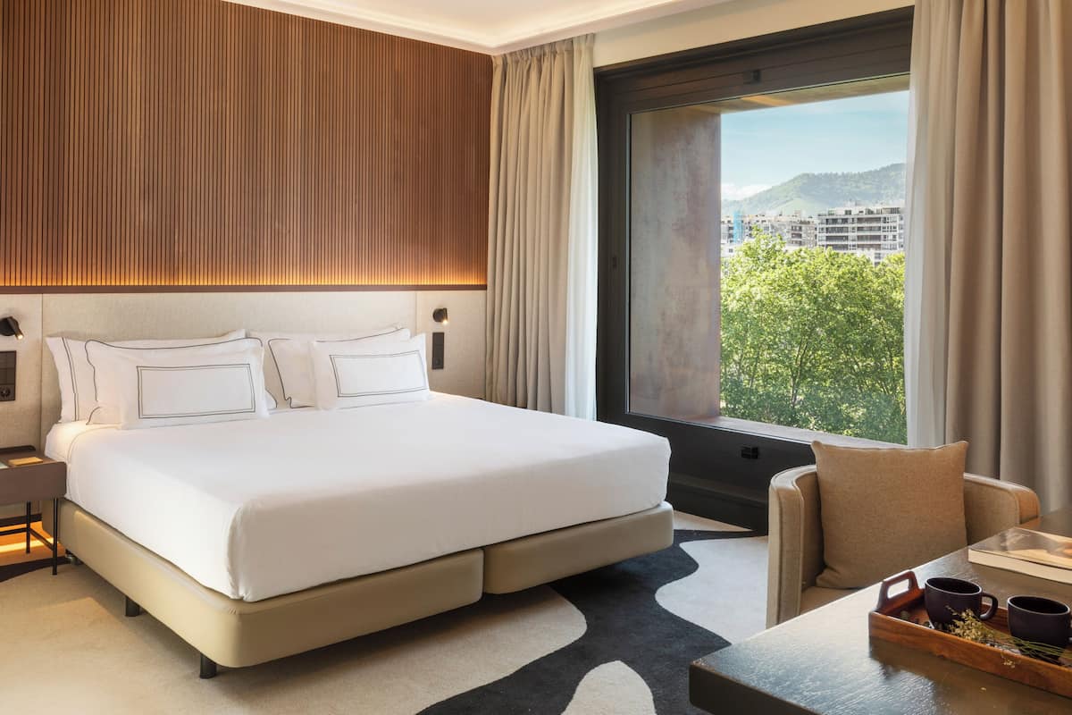 A modern hotel room with a large bed, a sofa, a dining table, and a view of a cityscape through a large window.