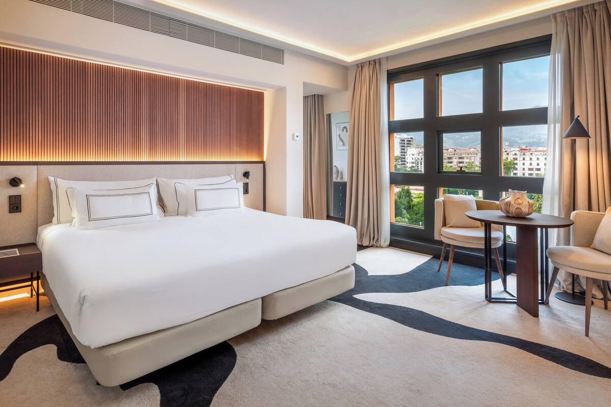 A modern hotel room with a large bed, a small table, and a view of the city through large windows.