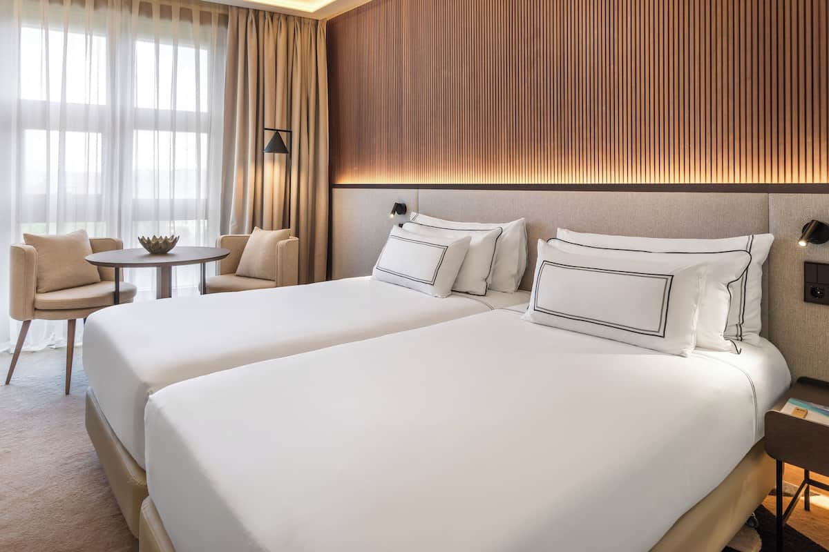 A hotel room with two beds, a small round table, a chair, and a view of the outdoors through sheer curtains.