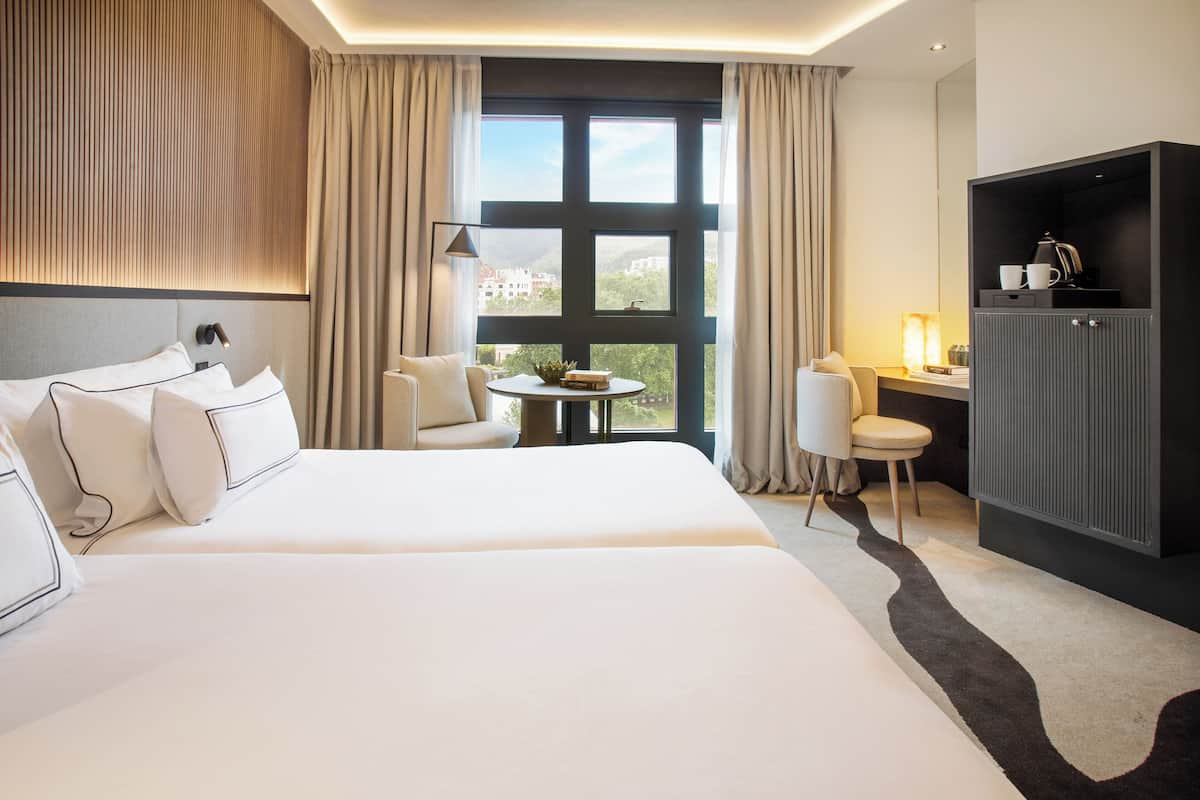 A hotel room with a large bed, a desk, a chair, and a view of the city through large windows.