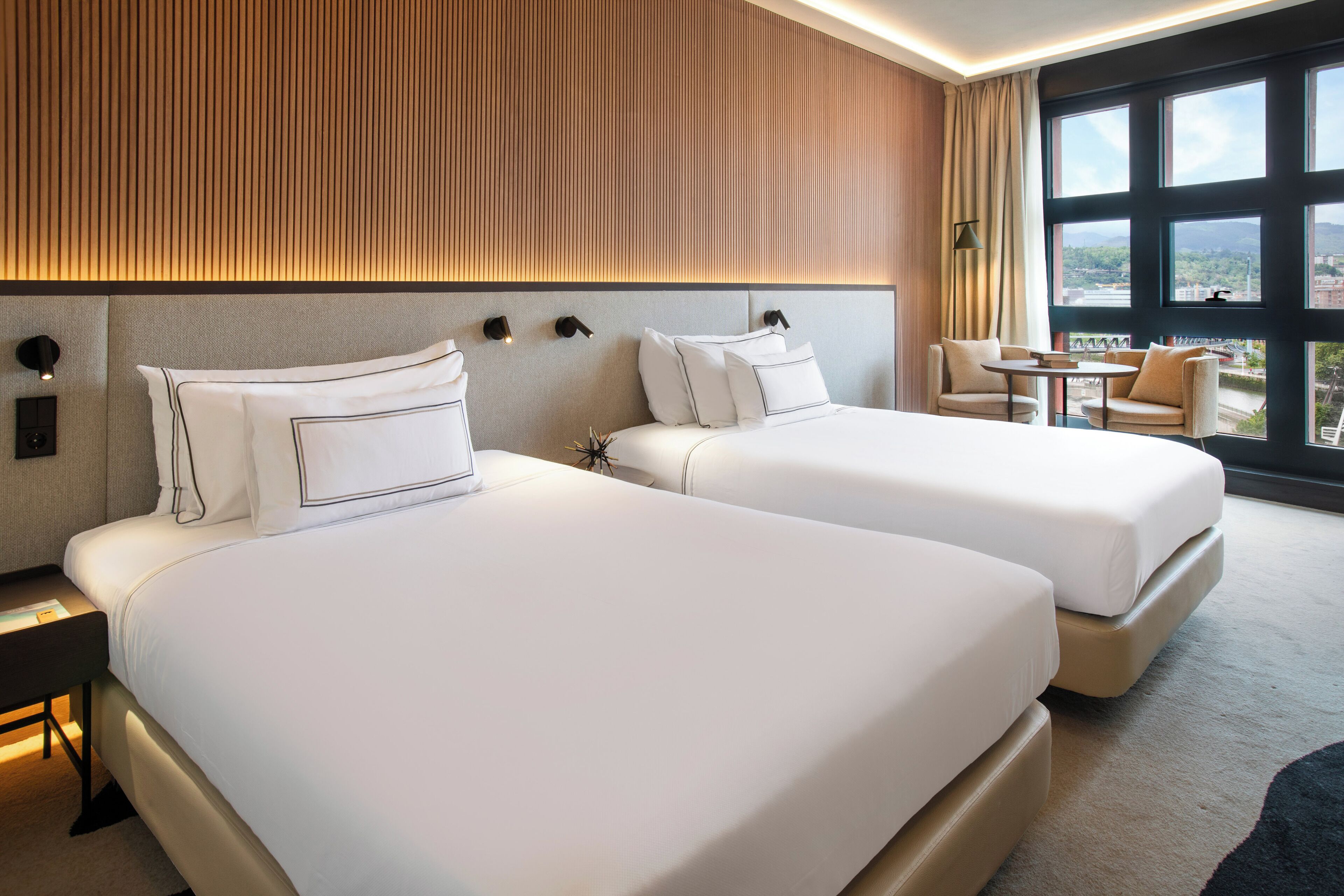 Habitación de hotel con dos camas, una mesita y vistas a la ciudad a través de amplios ventanales.