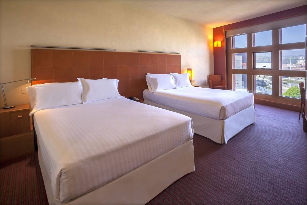 A hotel room with two beds, a large window with a view, and a nightstand with a lamp.