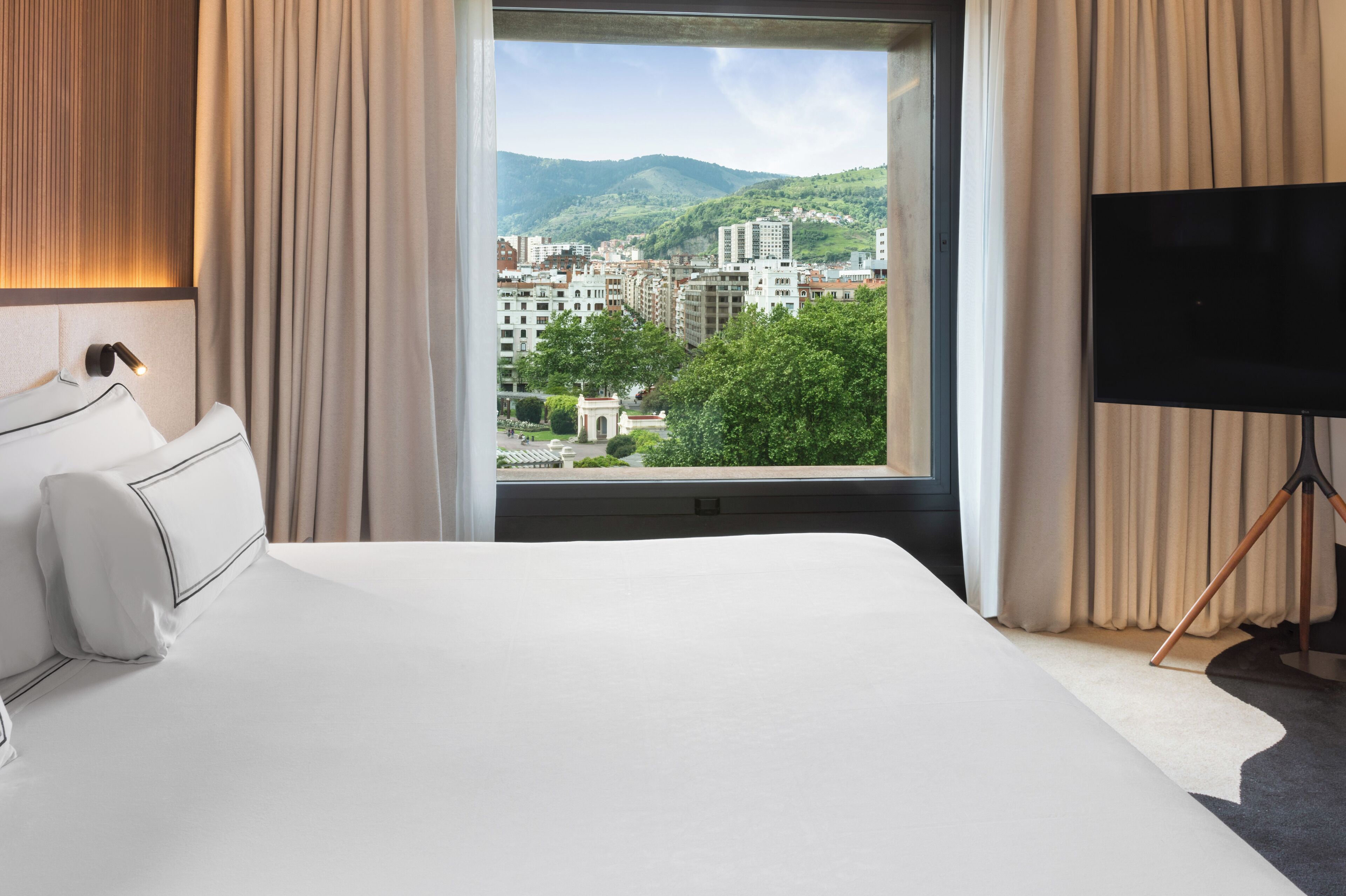 Habitación de hotel con una cama grande, un televisor sobre un soporte trípode y un ventanal con vistas a la ciudad.