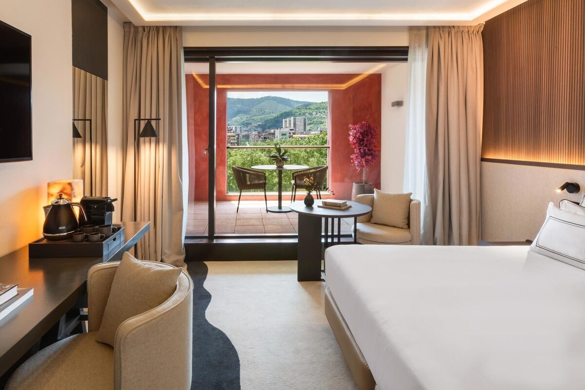 Habitación de hotel con cama, escritorio con cafetera, una silla, una mesita con un florero, y vistas a un balcón con mesa y sillas.