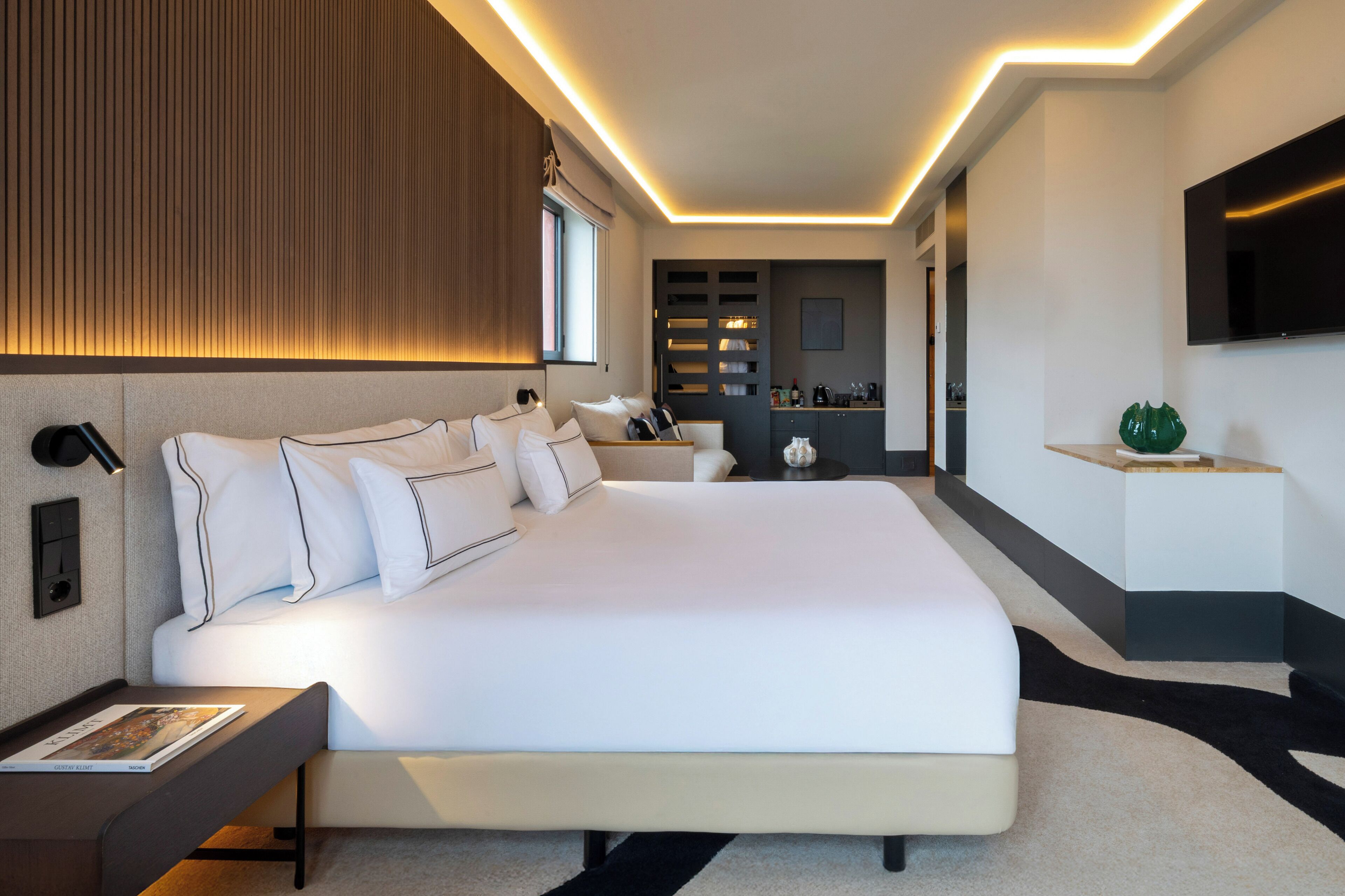 A modern hotel room with a large bed, bedside tables, a flat-screen TV, and a built-in kitchen area.