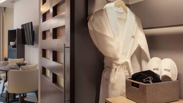 A hotel room with a modern design, featuring a wooden partition with cut-out panels, a seating area with a table and chairs, a robe hanging on a rack, and a basket containing slippers and a black item.