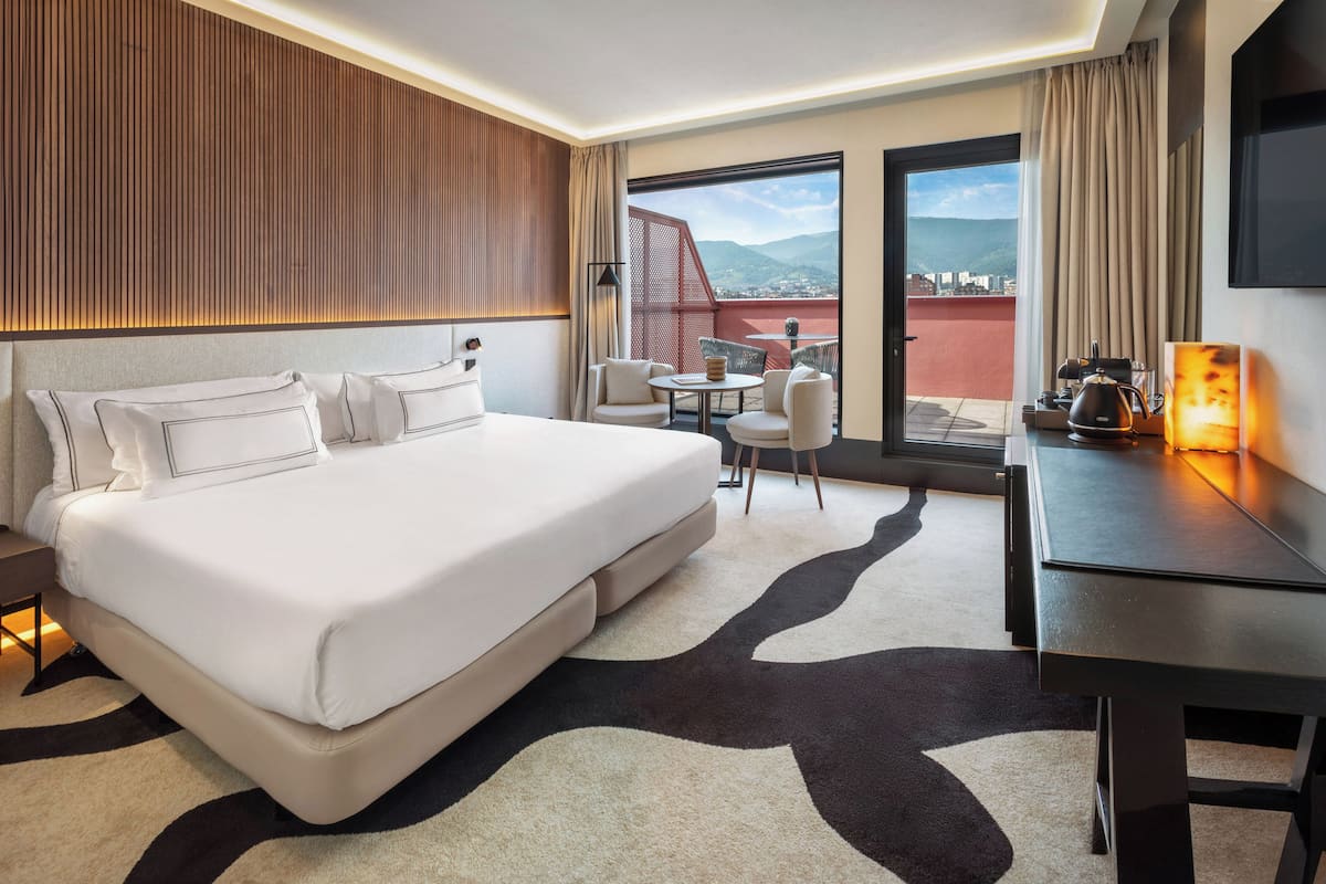 A hotel room with a large bed, a desk, a chair, a table, and a view of the city and mountains through the balcony.