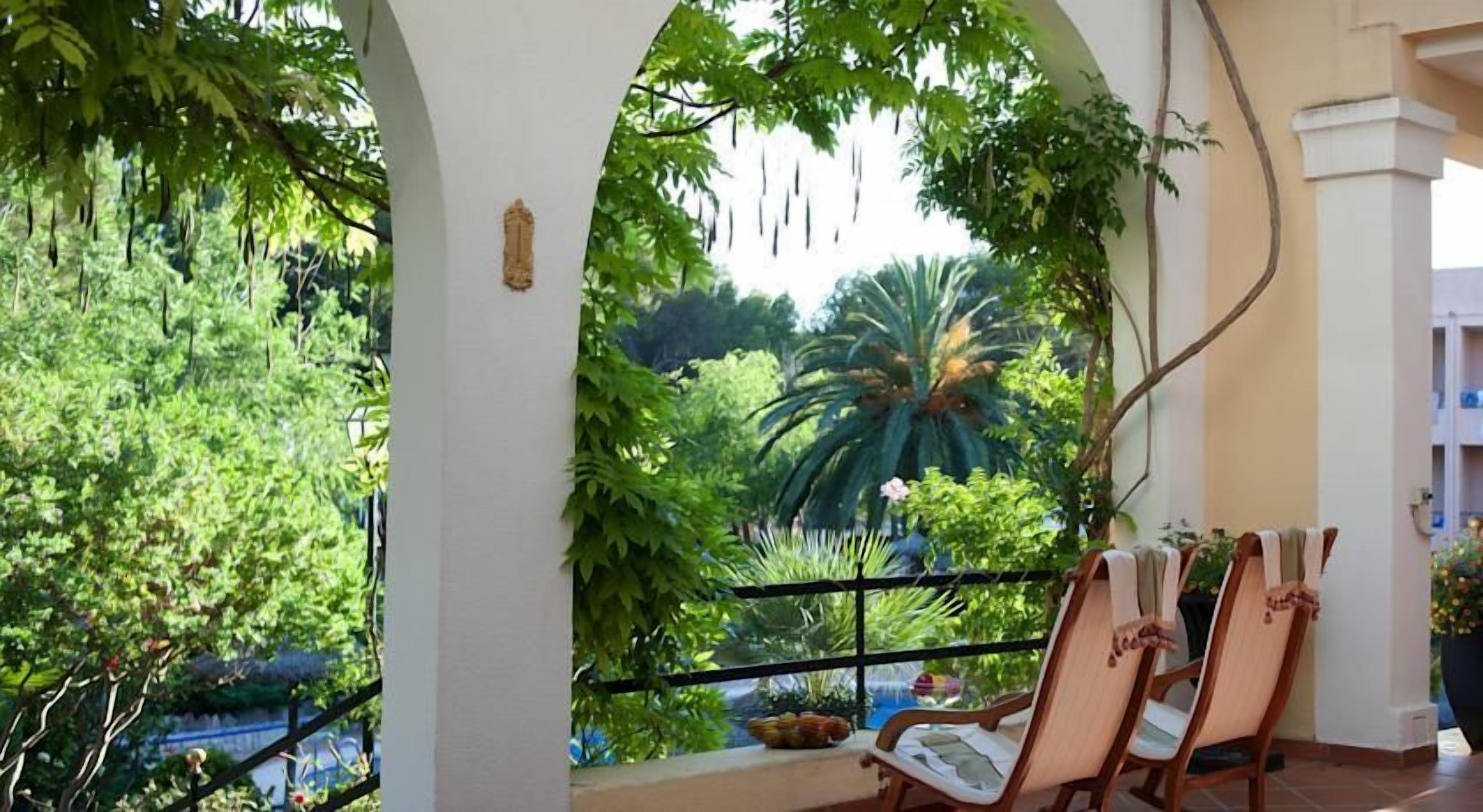 A balcony with chairs, a view of lush greenery, and a building in the background.