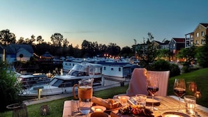 A dining area with a view of boats and houses, a picnic setup with food and drinks, and a grassy outdoor setting. - Precise Resort Marina Wolfsbruch (Rheinsberg)