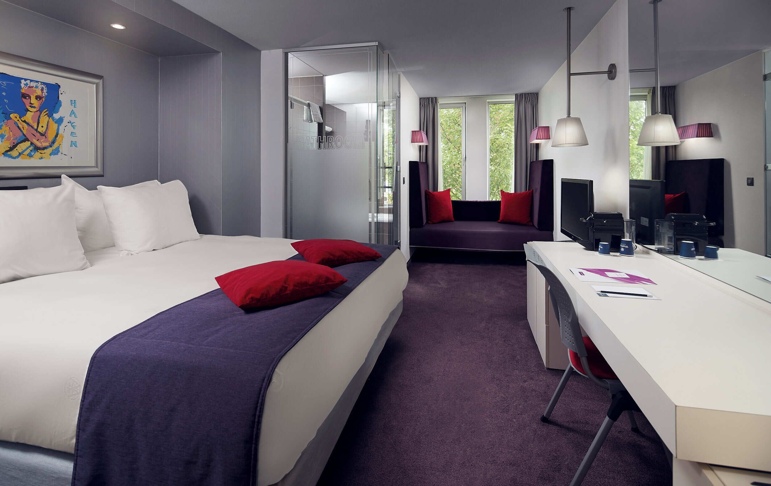 Ein modernes Hotelzimmer mit einem großen Bett, einem Schreibtisch mit Computer und einer Sitzecke mit Blick ins Freie.