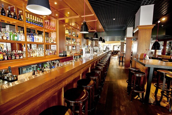 Un bar con estantes de madera llenos de botellas y vasos, varias lámparas colgantes y una barra larga con taburetes.
