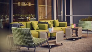 Un conjunto de sofá verde con sillones a juego y una pequeña mesa, ubicado frente a un edificio con un amplio ventanal que muestra el texto 'THE GALLERY'.