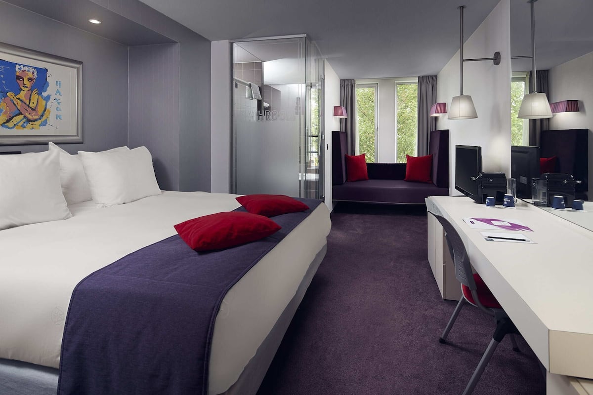 Una habitación de hotel moderna con una cama grande, un escritorio, una computadora y una ventana con vistas a árboles.