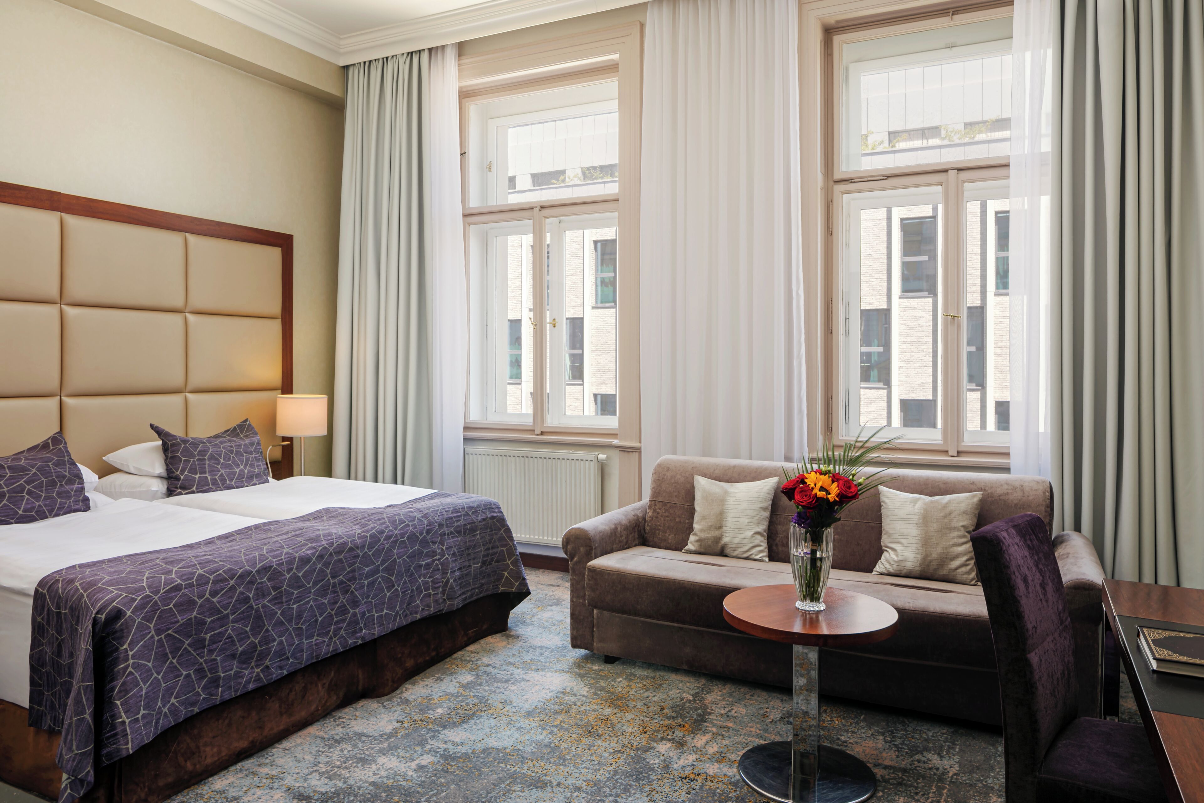 Une chambre d’hôtel avec un grand lit, un canapé, une petite table ornée d’un vase de fleurs et de grandes fenêtres avec des rideaux.