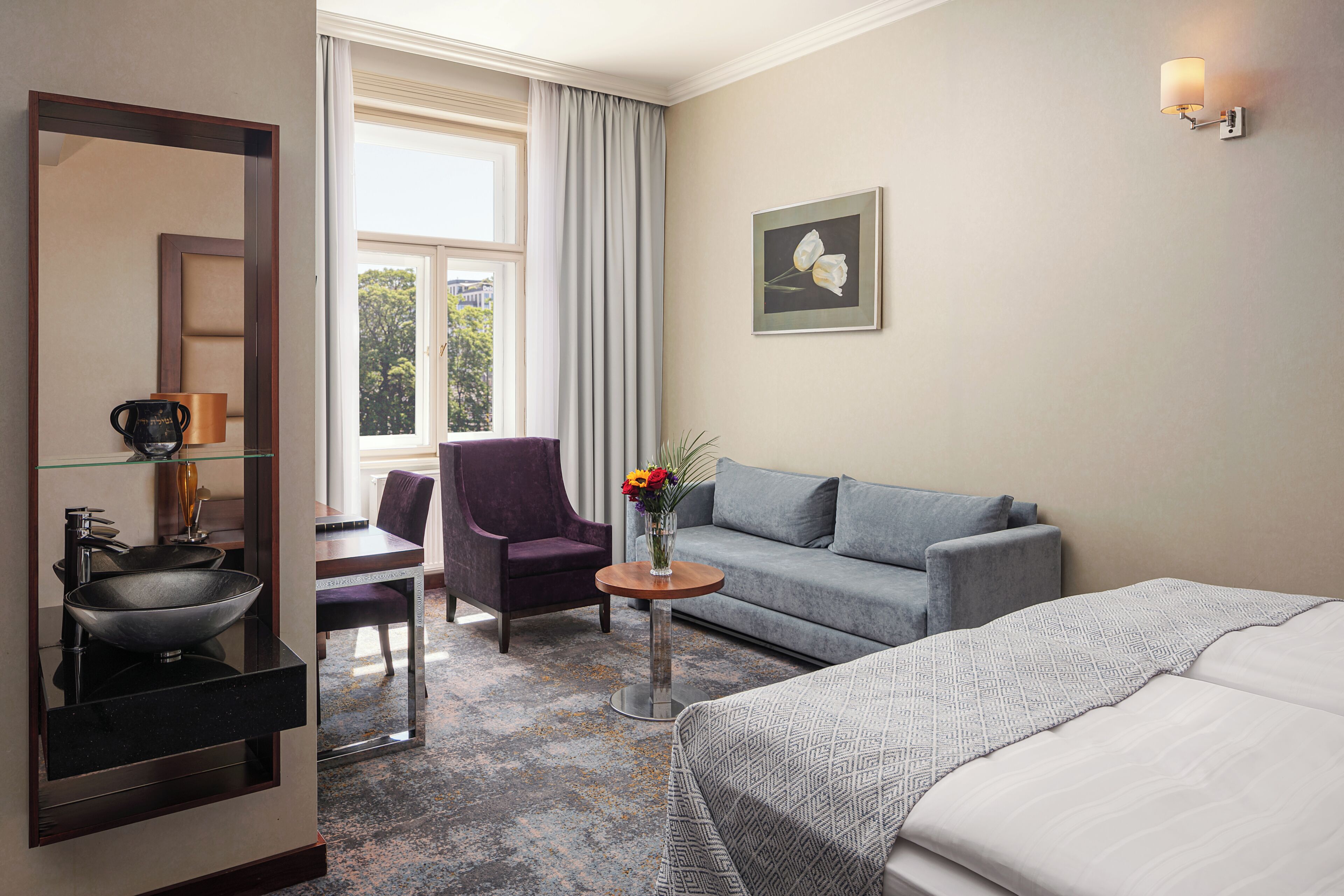Una camera d'albergo con un letto, una poltrona, una sedia, un tavolino con un vaso di fiori, uno specchio e una finestra con tende.