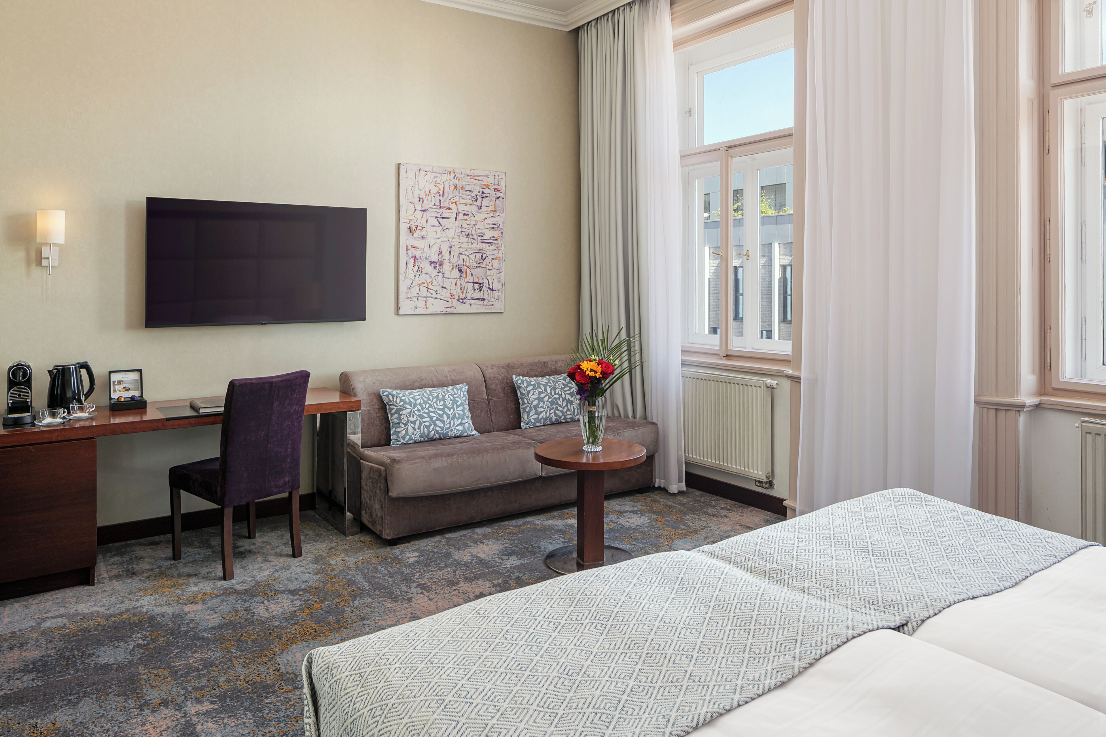 Habitación de hotel con cama, sofá, escritorio, silla, cafetera, un florero con flores, un cuadro y una ventana con cortinas.