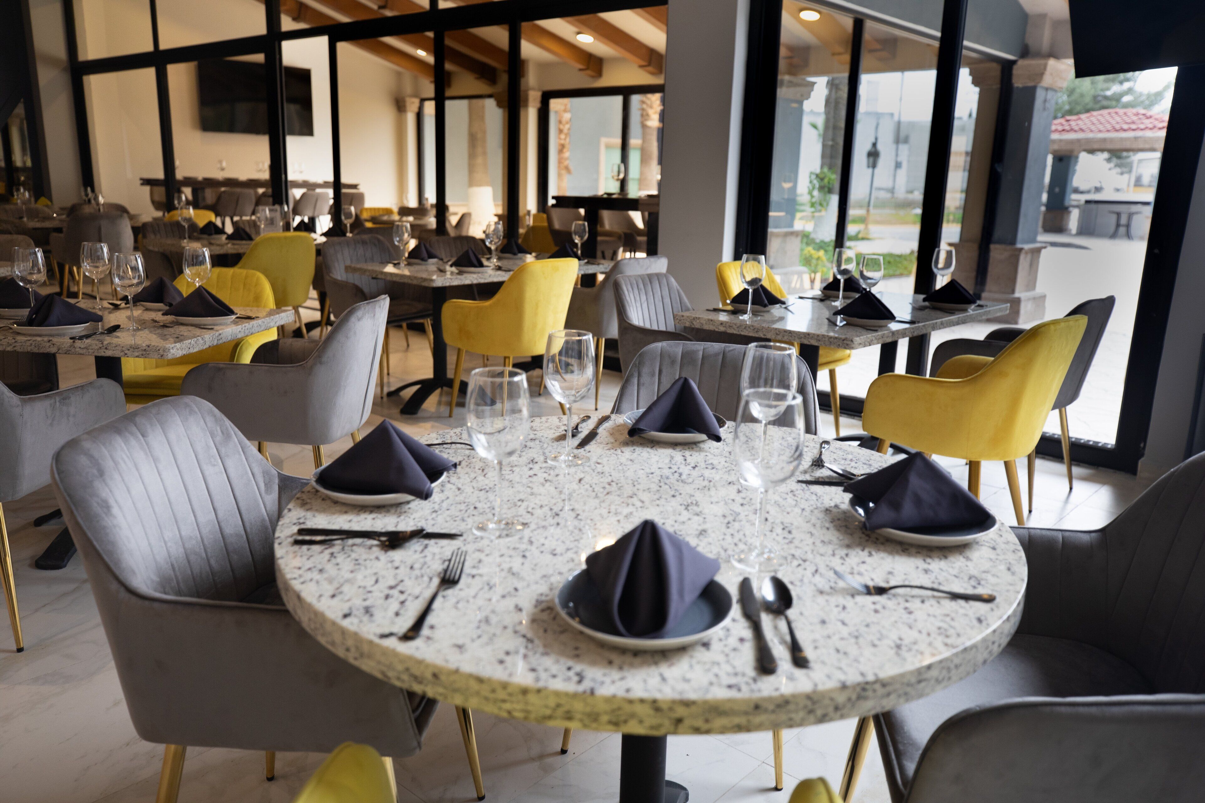 A restaurant with round tables, grey and yellow chairs, and large windows.
