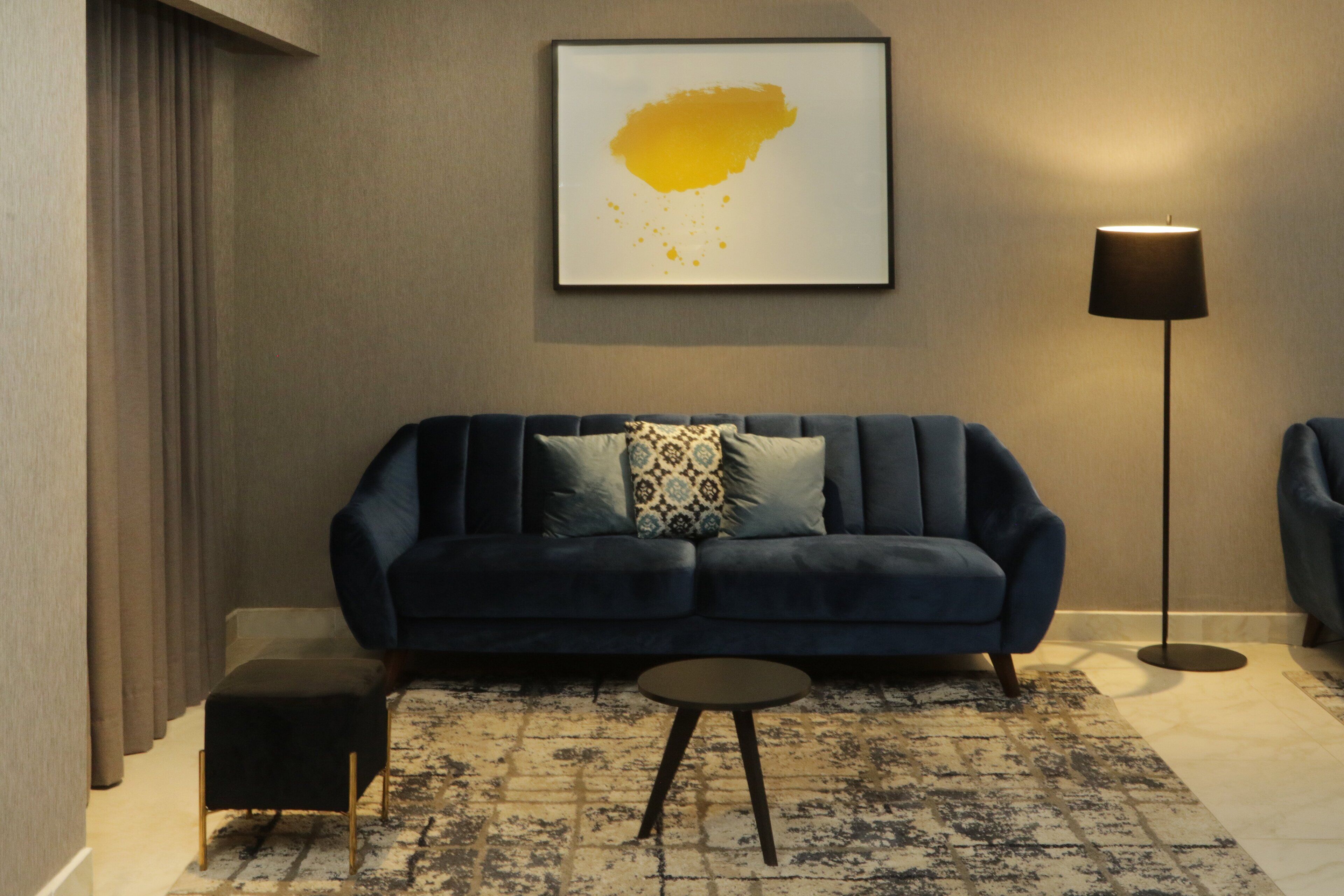 A modern living room with a dark blue sofa, a small round table, a floor lamp, and a framed artwork on the wall.