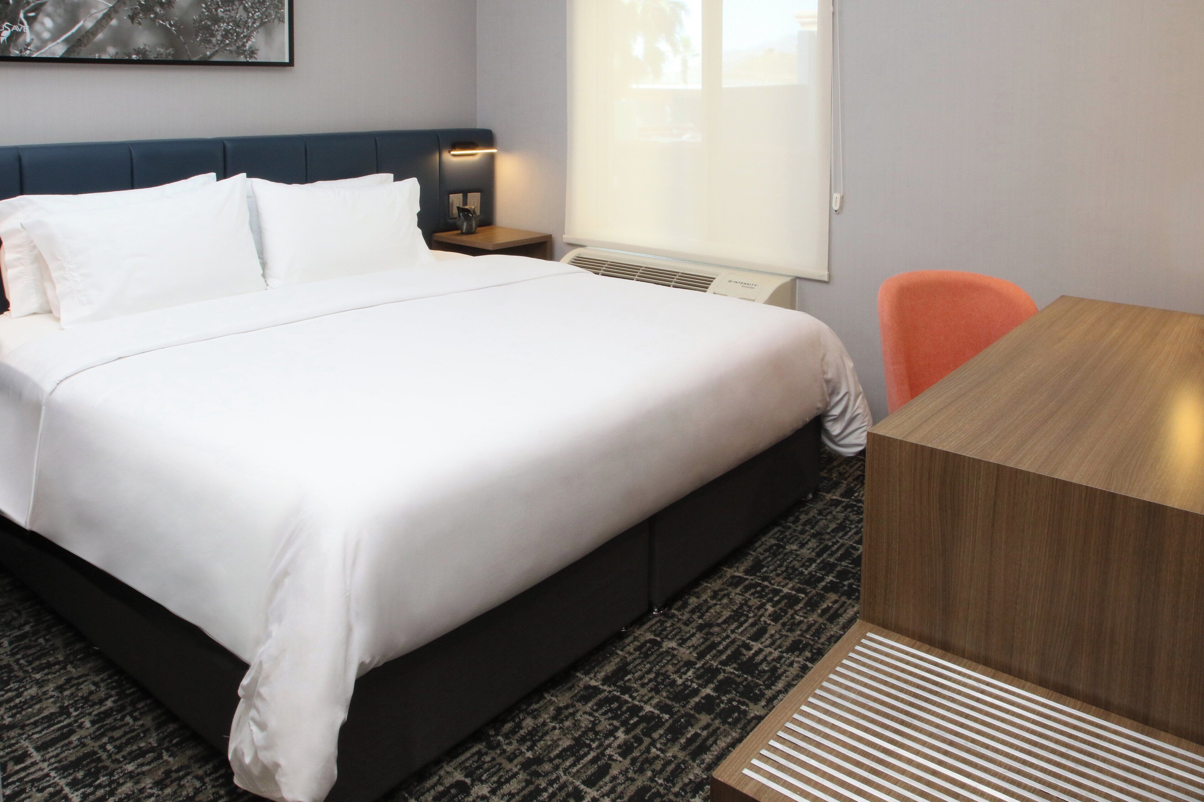 A hotel room with a large bed, a wooden desk, a nightstand, and a window with blinds.
