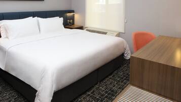 A hotel room with a large bed, a wooden desk, a nightstand, and a window with blinds.