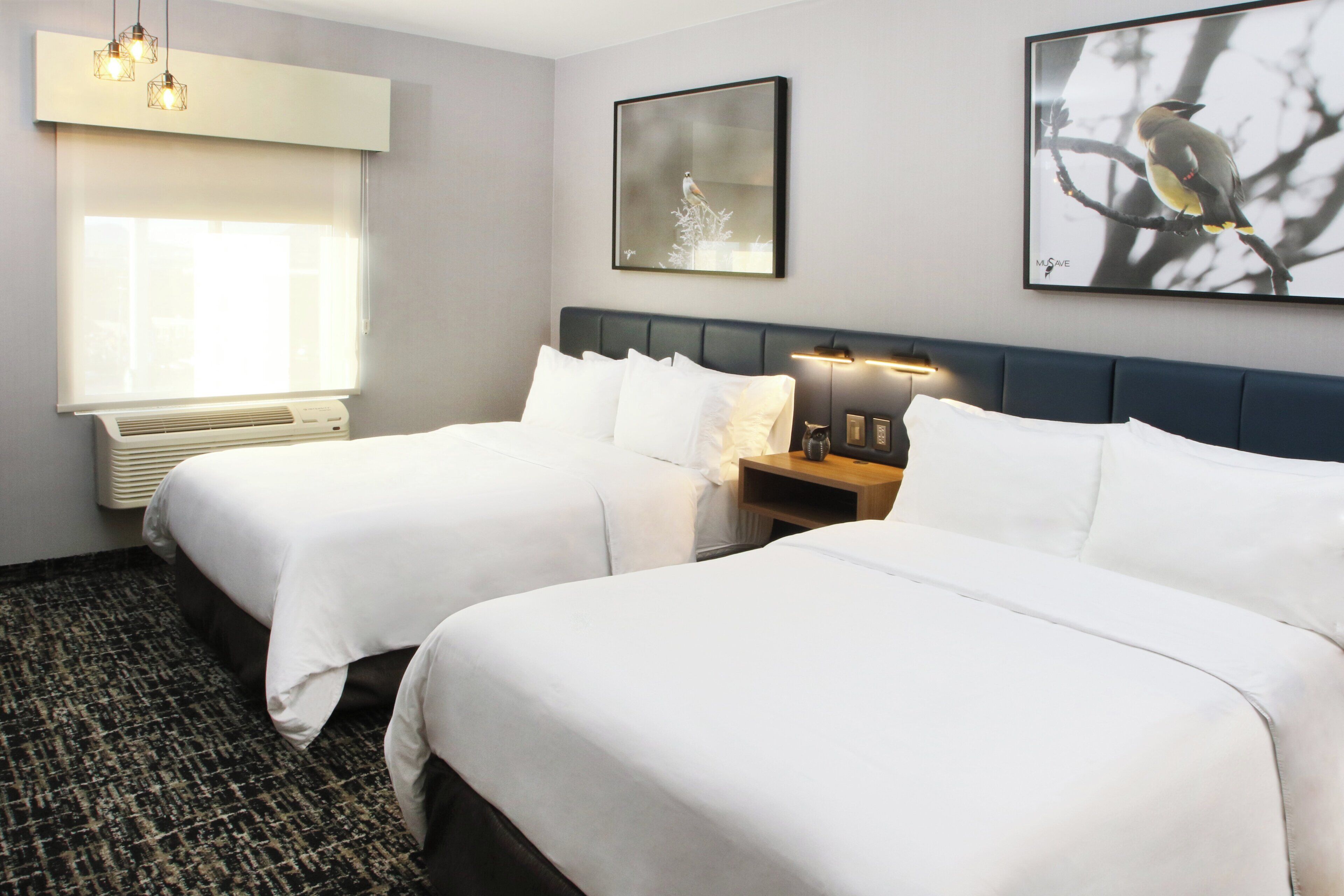 A hotel room with two beds, a window with blinds, a wall-mounted picture, and a nightstand with a lamp.