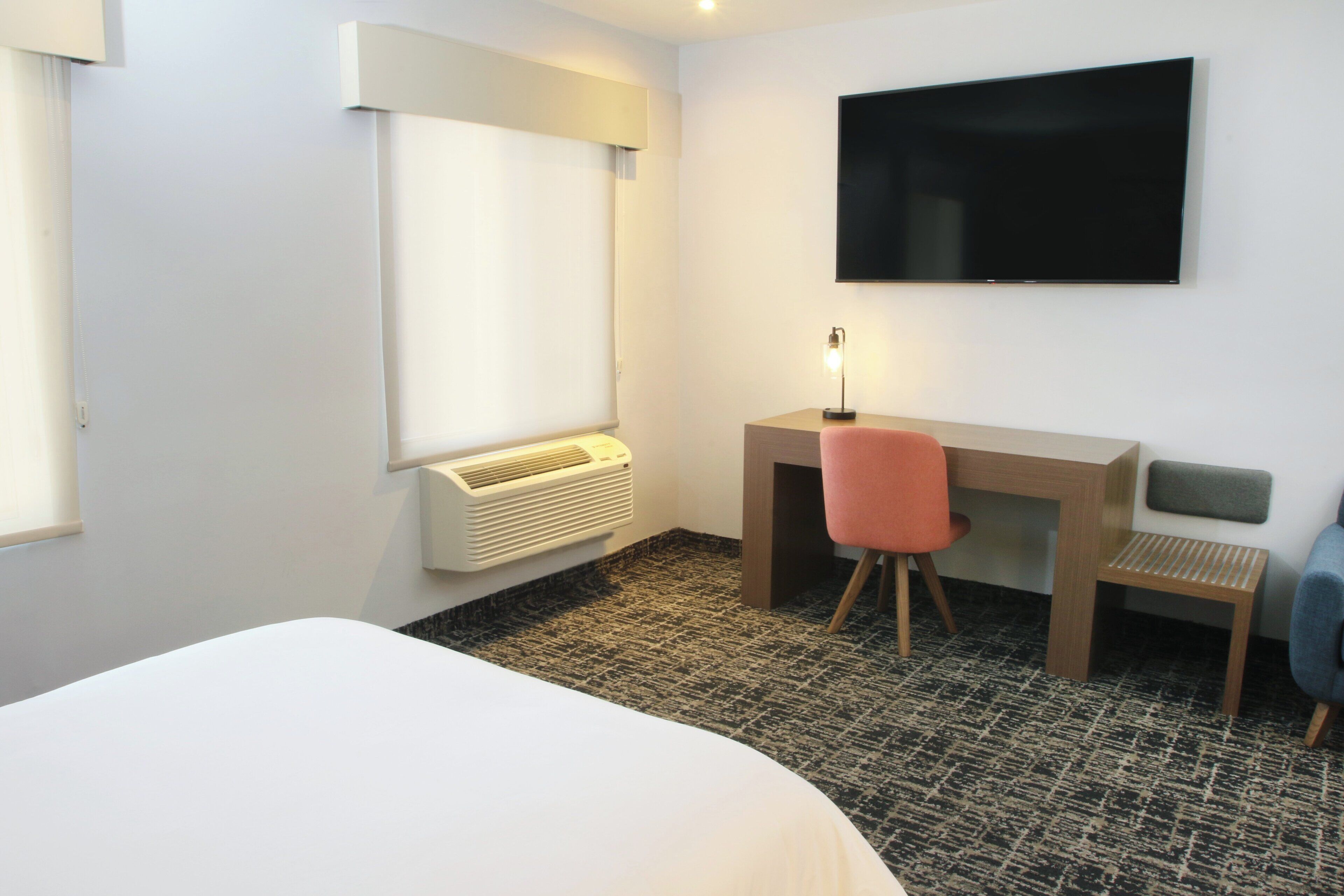 A hotel room with a bed, a desk with a chair, a television mounted on the wall, and a window with blinds.