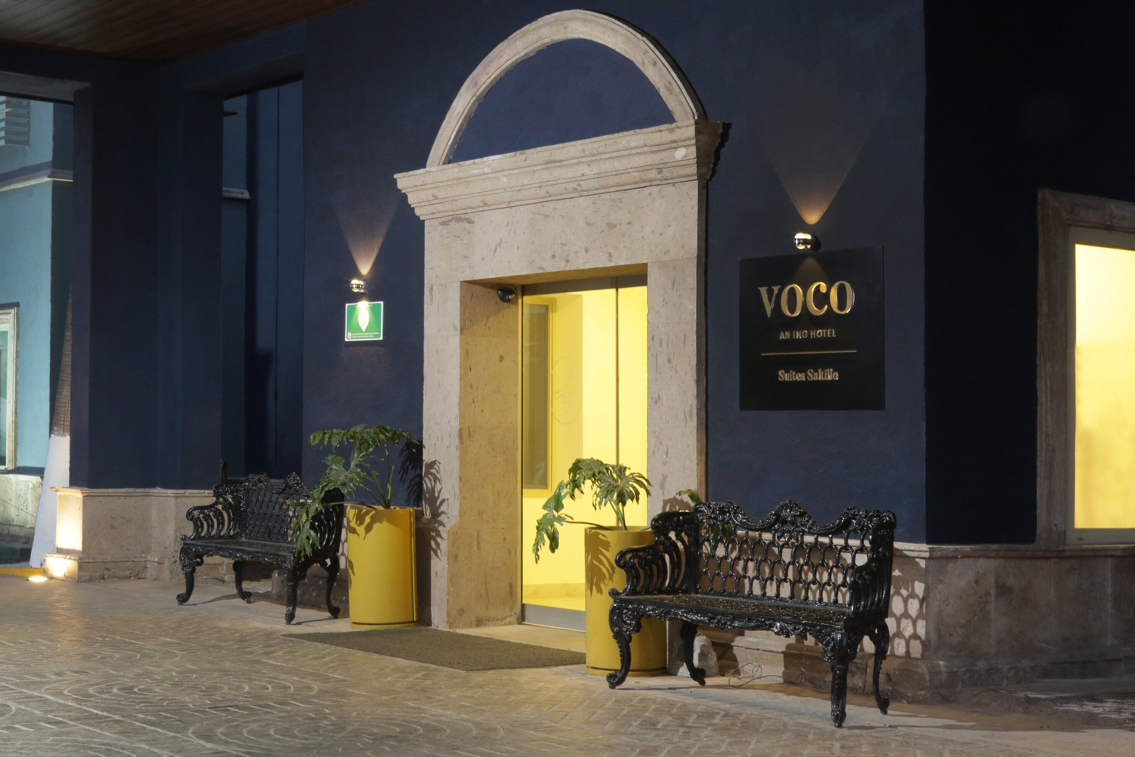 La entrada de un hotel con un cartel que dice "VOCO AN INGO HOTEL Suites Salinas". Hay dos bancos fuera de la entrada, y se ve una maceta a la izquierda.