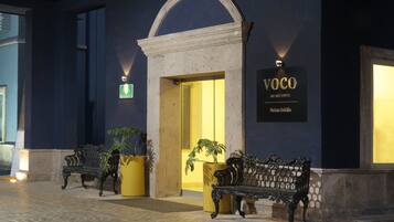 La entrada de un hotel con un cartel que dice "VOCO AN INGO HOTEL Suites Salinas". Hay dos bancos fuera de la entrada, y se ve una maceta a la izquierda.