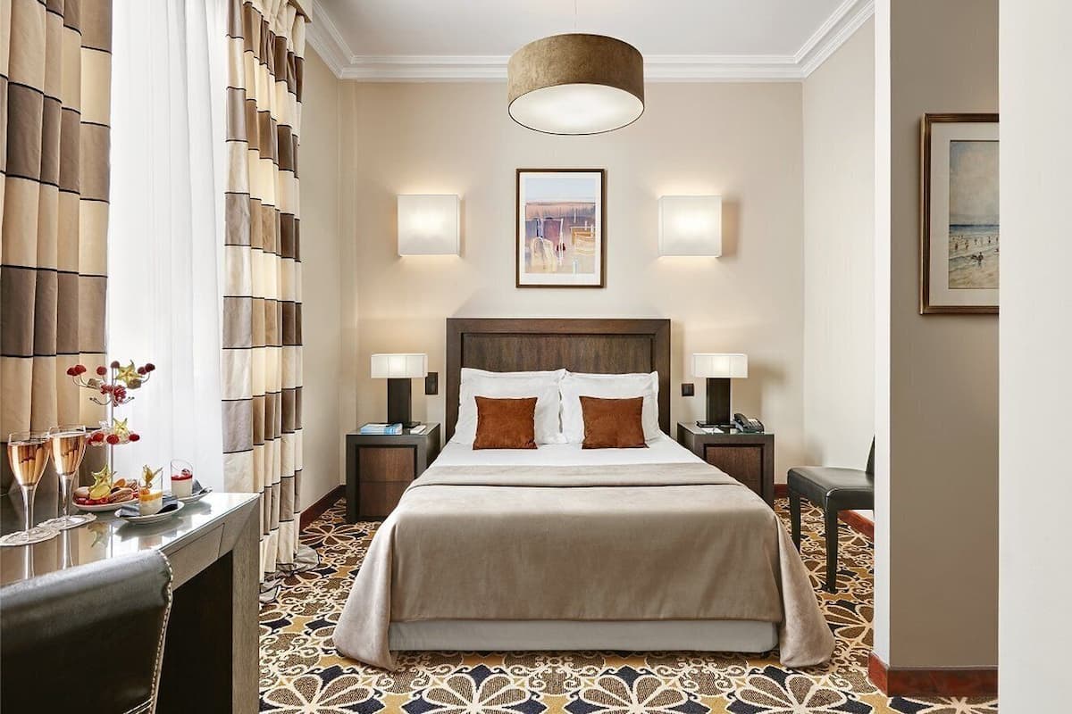 Habitación de hotel con una cama grande, dos mesitas de noche, un escritorio con una silla, alfombra estampada y una lámpara en el techo.