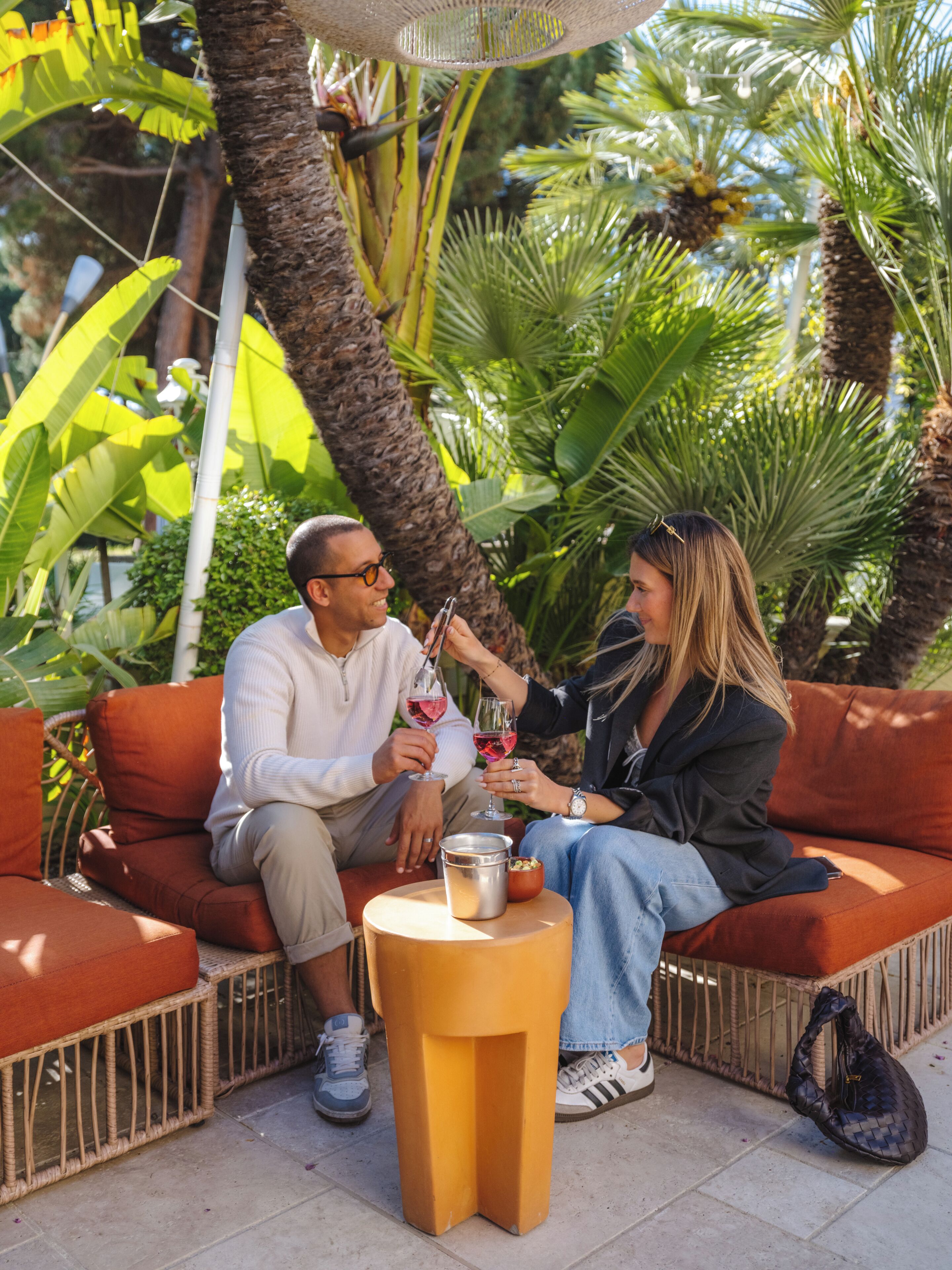 Un couple est assis sur un canapé en osier, devant une petite table ronde, le tout étant entouré d’une végétation luxuriante.