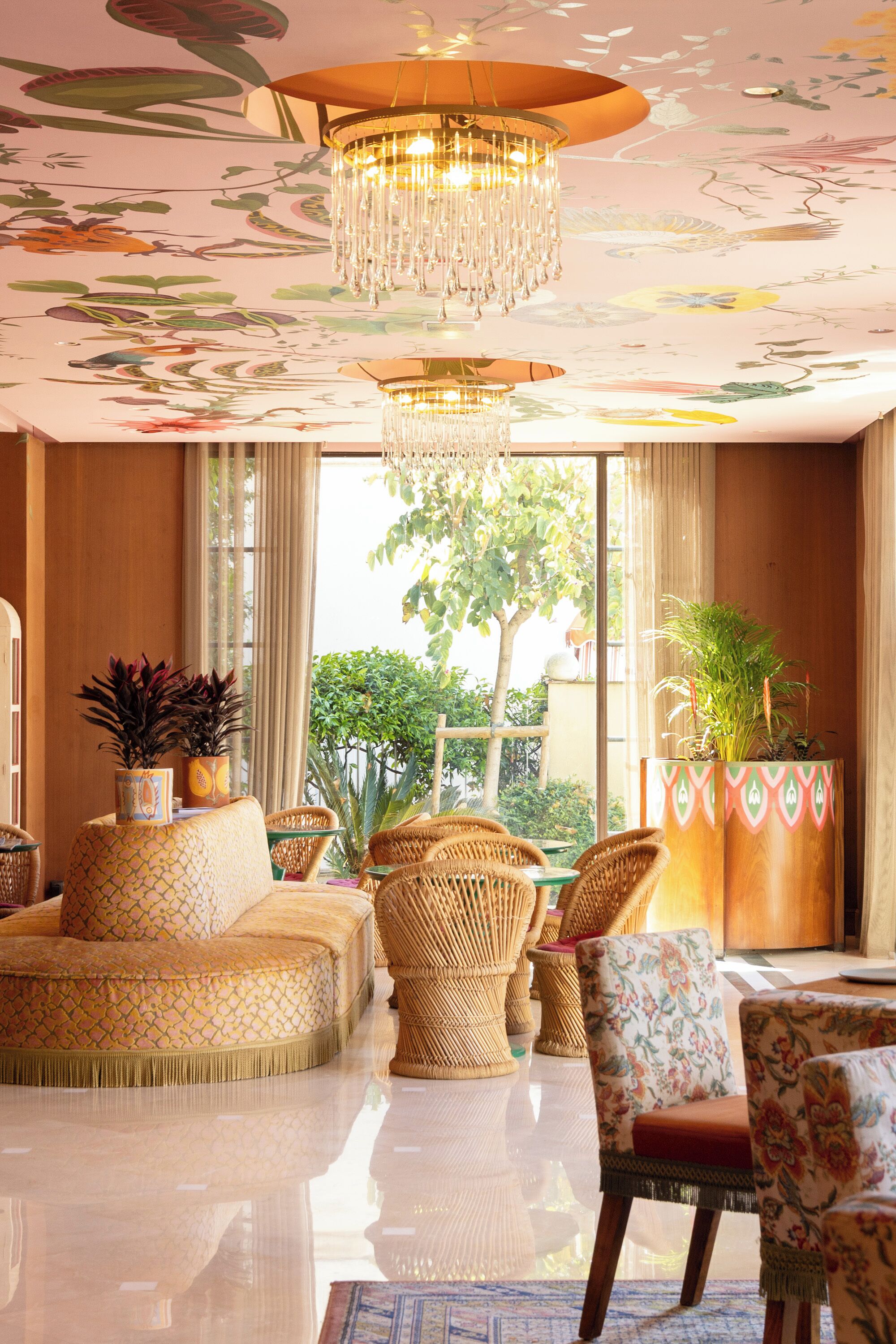 A spacious living room with a large chandelier, wicker furniture, and a view of greenery outside.